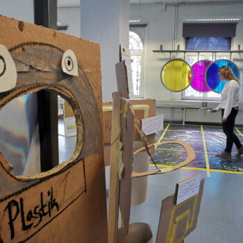 Kunstinstallation mit einem braunen Pappaufsteller mit ausgeschnittenem Mund und zwei weißen Augen, im Hintergrund eine Person vor drei großen, farbigen Kreisen an der Wand in einem hellen Raum mit großen Fenstern.