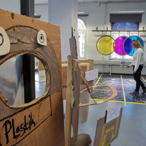 Kunstinstallation mit einem braunen Pappaufsteller mit ausgeschnittenem Mund und zwei weißen Augen, im Hintergrund eine Person vor drei großen, farbigen Kreisen an der Wand in einem hellen Raum mit großen Fenstern.
