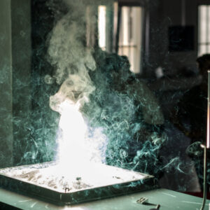 Dichter weißer Rauch steigt aus einem flachen Behälter auf einem Tisch in einem dunklen Raum mit Fenstern im Hintergrund.