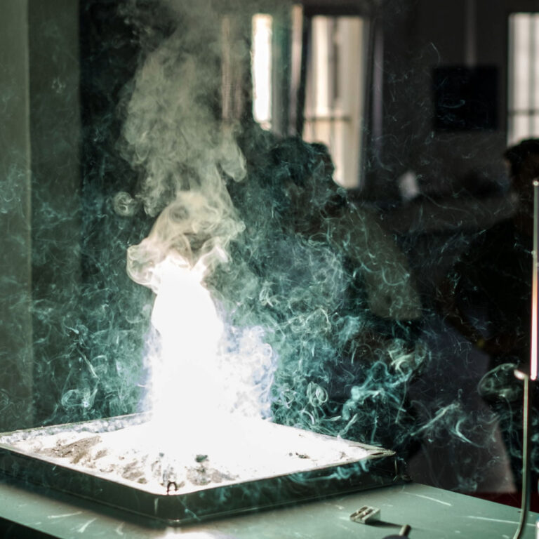 Dichter weißer Rauch steigt aus einem flachen Behälter auf einem Tisch in einem dunklen Raum mit Fenstern im Hintergrund.