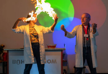 Zwei Personen in weißen Laborkitteln führen eine wissenschaftliche Vorführung durch, bei der eine Flamme über den Händen einer Person aufsteigt, bunte Lichtkreise sind an der Wand zu sehen.