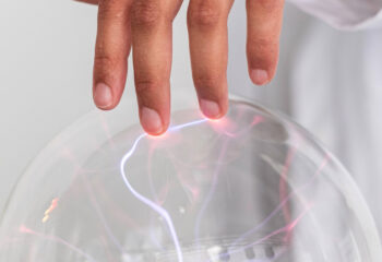 Finger berührt eine leuchtende Plasmakugel, aus der violette und rote elektrische Entladungen strahlen.