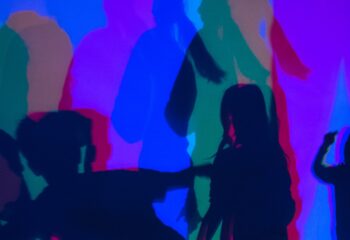 Mehrere farbige Schatten von Personen auf einer Wand, überwiegend in Blau, Rot und Grün.