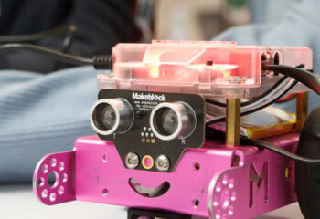 Kleiner pinkfarbener Roboter mit zwei runden Sensoren vorne, die wie Augen aussehen, und einem leuchtend roten Gehäuse oben.