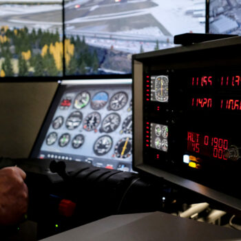 Cockpit eines Flugsimulators mit Instrumententafel, Steuerknüppel und digitaler Anzeige, im Hintergrund Flughafen und Landebahn sichtbar.