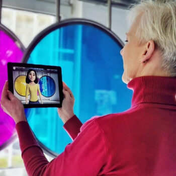 Person mit rotem Rollkragenpullover hält ein Tablet, auf dem eine animierte Figur in gelbem Oberteil vor bunten runden Fenstern zu sehen ist.