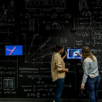 Zwei Personen vor einer Tafel mit mathematischen Formeln und Diagrammen, eine zeigt auf einen Bildschirm mit einer technischen Zeichnung.