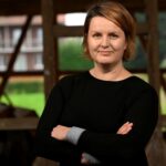 Frau mit kurzen, rotbraunen Haaren trägt schwarzen Pullover und steht mit verschränkten Armen vor einem unscharfem Hintergrund.