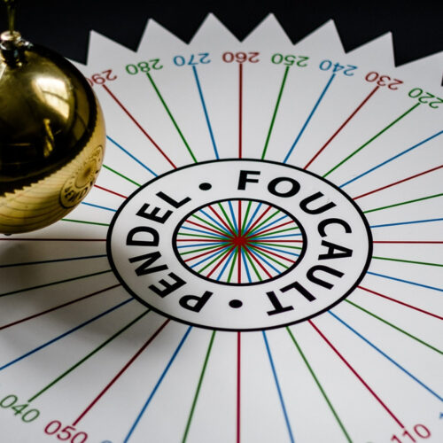 Pendelkugel über einer kreisförmigen Skala mit dem Text 'Pendel Foucault' und farbigen Linien in radialer Anordnung.