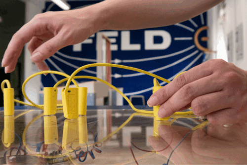 Hände greifen gelbe Kunststoffteile mit Drähten auf einem Tisch, im Hintergrund unscharf das Wort 'FELD' auf blauem Grund.
