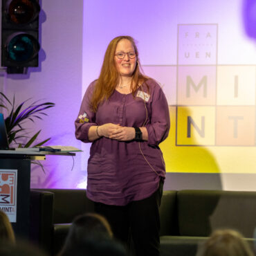 Frau steht vor Leinwand mit Präsentation, Publikum sitzt auf Stühlen in einem Veranstaltungsraum mit Beleuchtung und Lautsprechern.