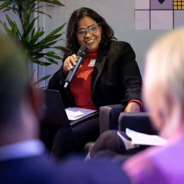 Frau sitzt Publikum sitzt auf Stühlen in einem Veranstaltungsraum mit Beleuchtung und Lautsprechern.