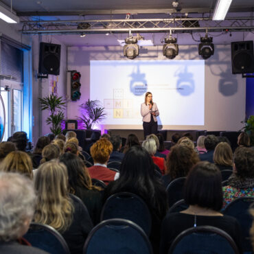 Frau steht vor Leinwand mit Präsentation, Publikum sitzt auf Stühlen in einem Veranstaltungsraum mit Beleuchtung und Lautsprechern.