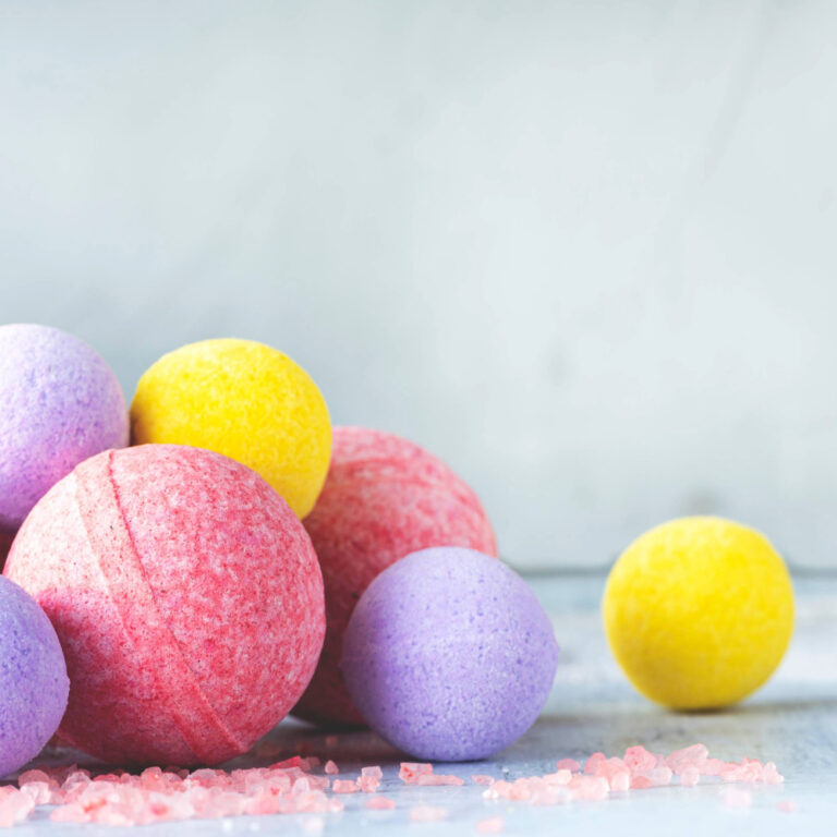 Mehrere bunte Badebomben in Rosa, Gelb und Lila liegen auf einer Oberfläche mit rosa Badesalz verstreut.