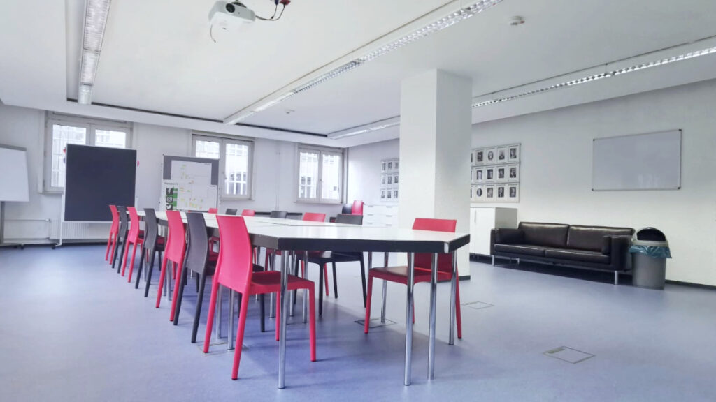 Konferenzraum mit langen Tischen, roten Stühlen, schwarzem Sofa und mehreren Fenstern.