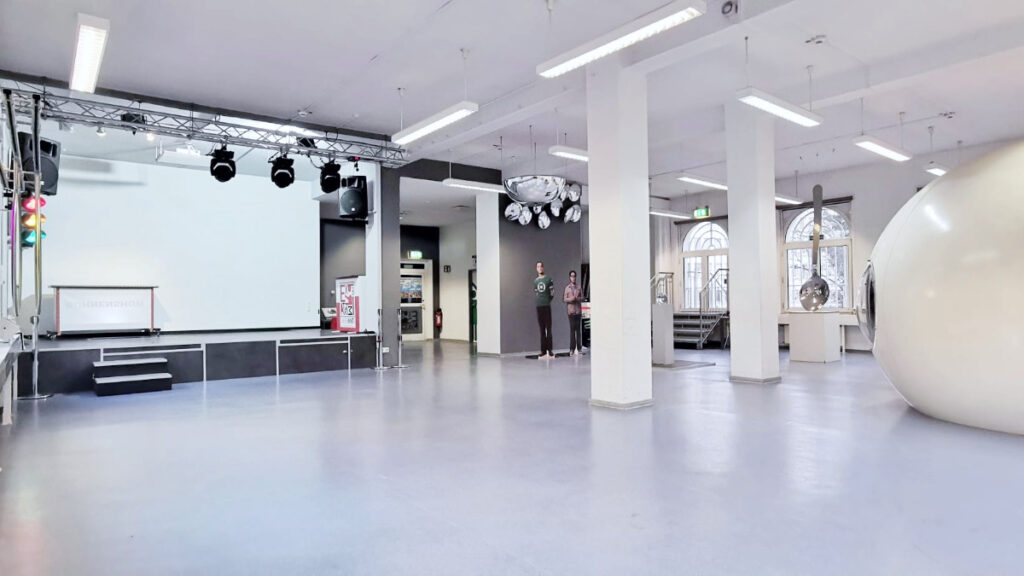 Leerer heller Raum mit Bühne, Treppenstufen und drei farbigen, runden Scheiben an der Fensterfront.