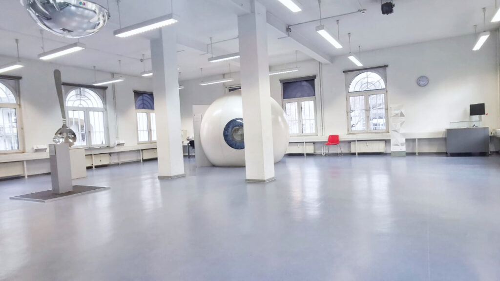 Großer, leerer Raum mit grauem Boden, weißen Wänden und hohen Fenstern, darin zwei große, runde Skulpturen und ein roter Stuhl.