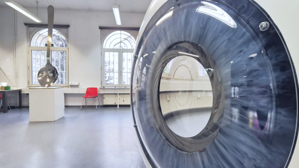 Großes, rundes Gerät mit transparentem Ring in hellem Raum mit Fenstern und rotem Stuhl.