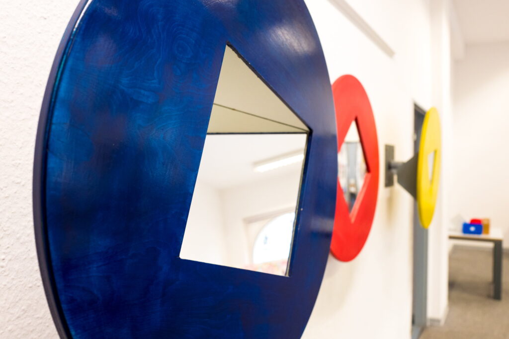 Drei runde Spiegel mit quadratischer Aussparung in den Farben Blau, Rot und Gelb an einer weißen Wand.