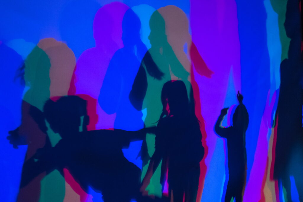 Mehrere farbige Schatten von Personen auf einer Wand, überwiegend in Blau, Rot und Grün.