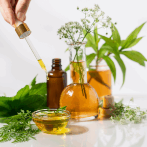 Hand hält Pipette mit gelblicher Flüssigkeit über kleine Glasschale, umgeben von grünen Blättern und Glasgefäßen mit Pflanzen und Flüssigkeiten.