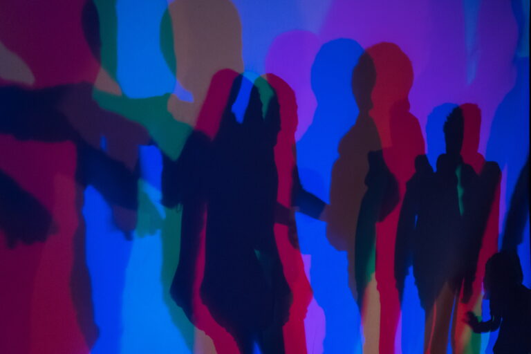 Mehrere farbige Schatten von Personen auf einer Wand, überwiegend in Blau, Rot und Grün.