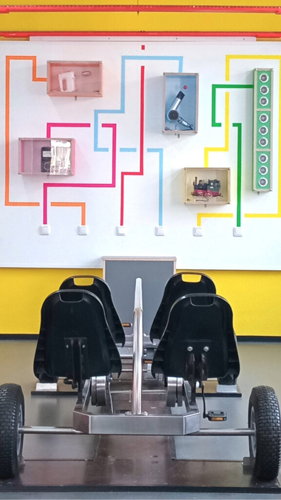 Zwei schwarze Sitze auf einem Metallgestell mit vier Reifen vor einer Wand mit farbigen Leitungen, die zu verschiedenen technischen Geräten führen.