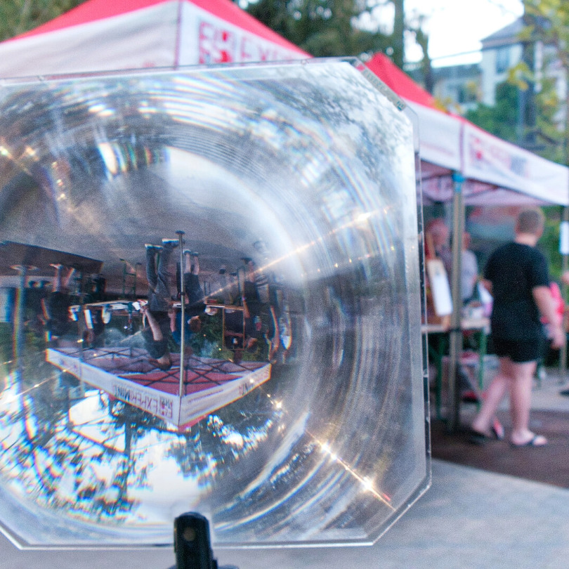 Vergrößernde Linse zeigt auf einem Marktstand eine umgekehrte Spiegelung von Personen und einem Zelt mit rotem Dach.