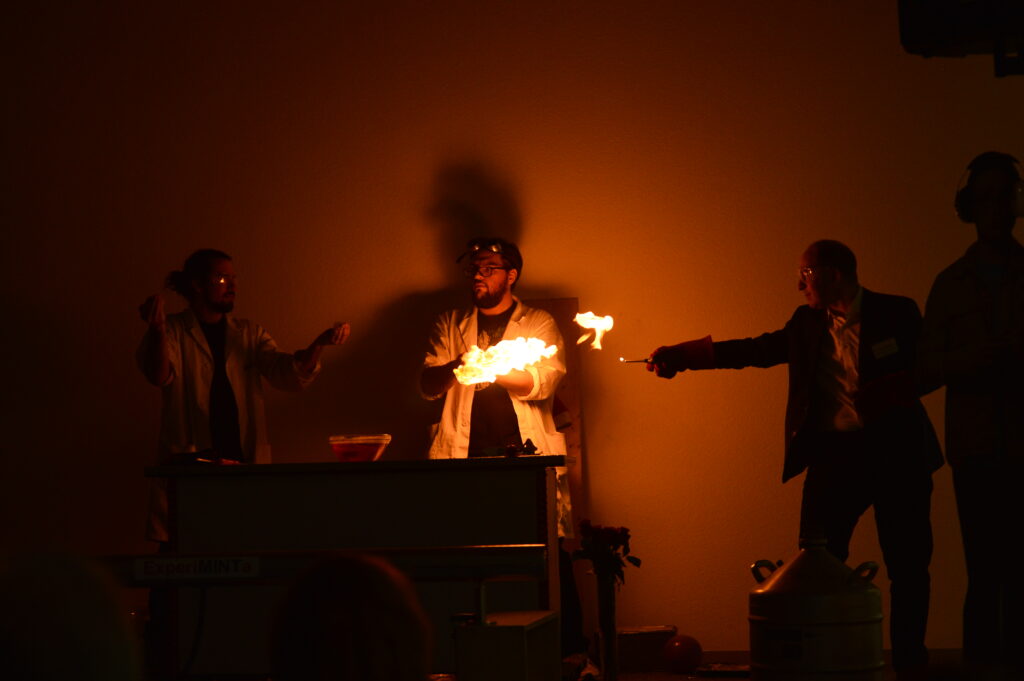 Drei Personen in einem dunklen Raum führen ein Experiment mit einer sichtbaren Flamme durch, die von der Mitte nach rechts leuchtet.