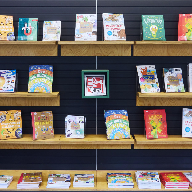 Regal mit mehreren bunten Kinderbüchern zu Wissenschaftsthemen, mittig ein grüner Rahmen mit dem Schriftzug 'EXPERIMINTA' in rot und schwarz.