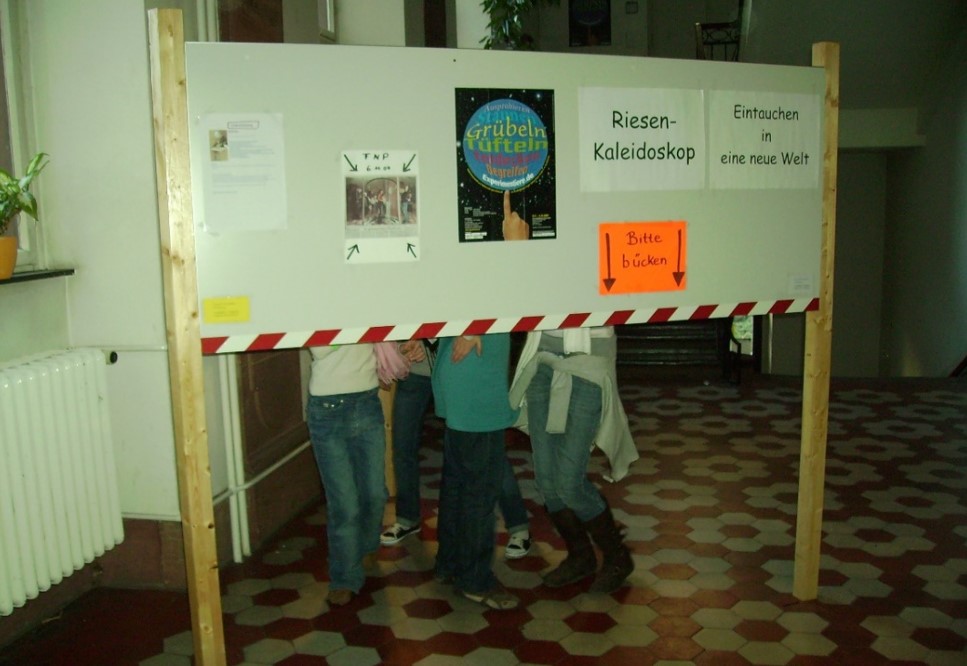 Vier Personen stehen unter einem großen weißen Brett mit Texten und Bildern, darunter 'Riesen-Kaleidoskop' und 'Bitte bücken' auf einem orangefarbenen Schild.